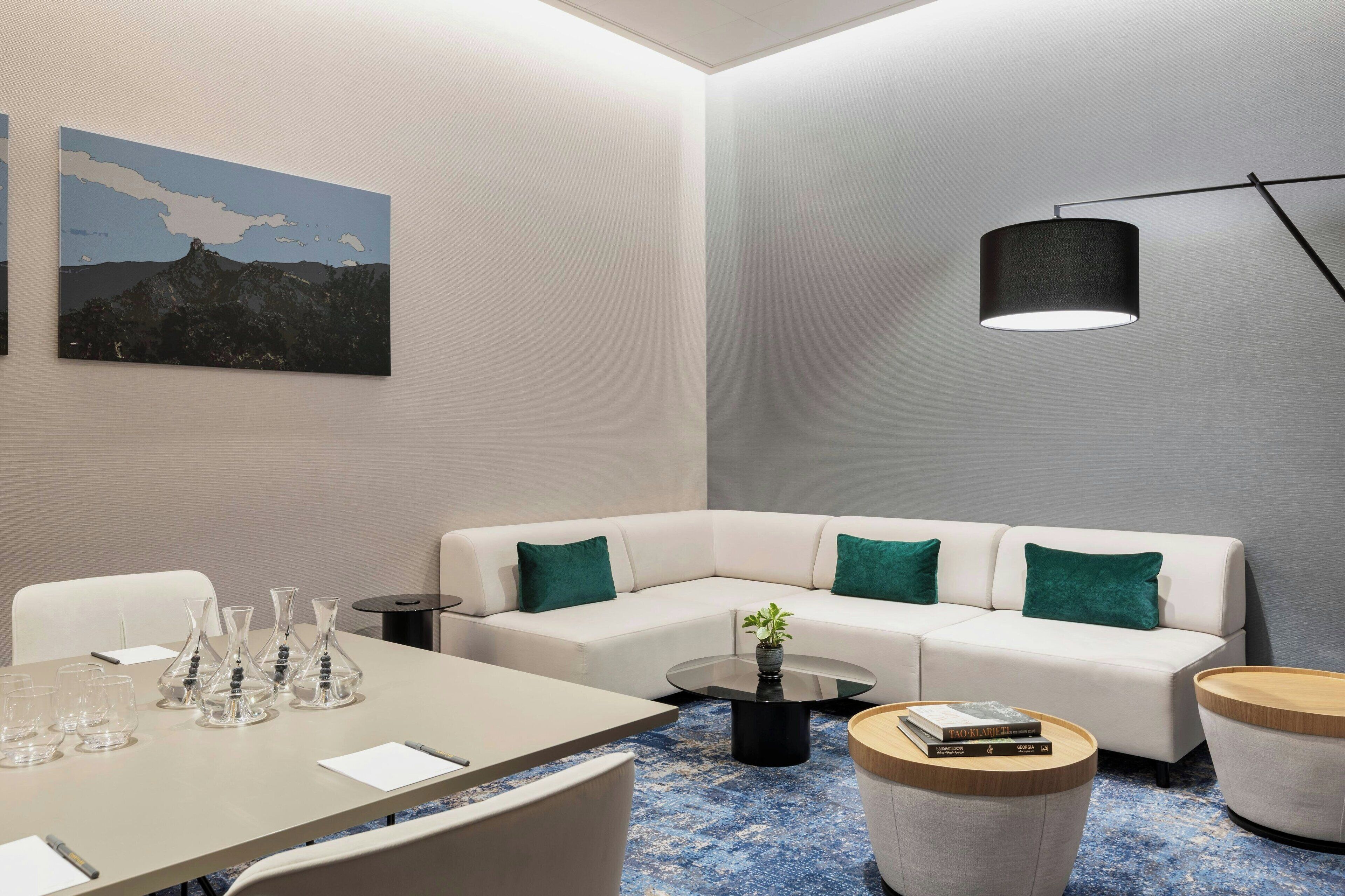A modern hotel room with a white sofa, a round coffee table, and a dining table with chairs. There is a painting on the wall and a lamp on the ceiling.