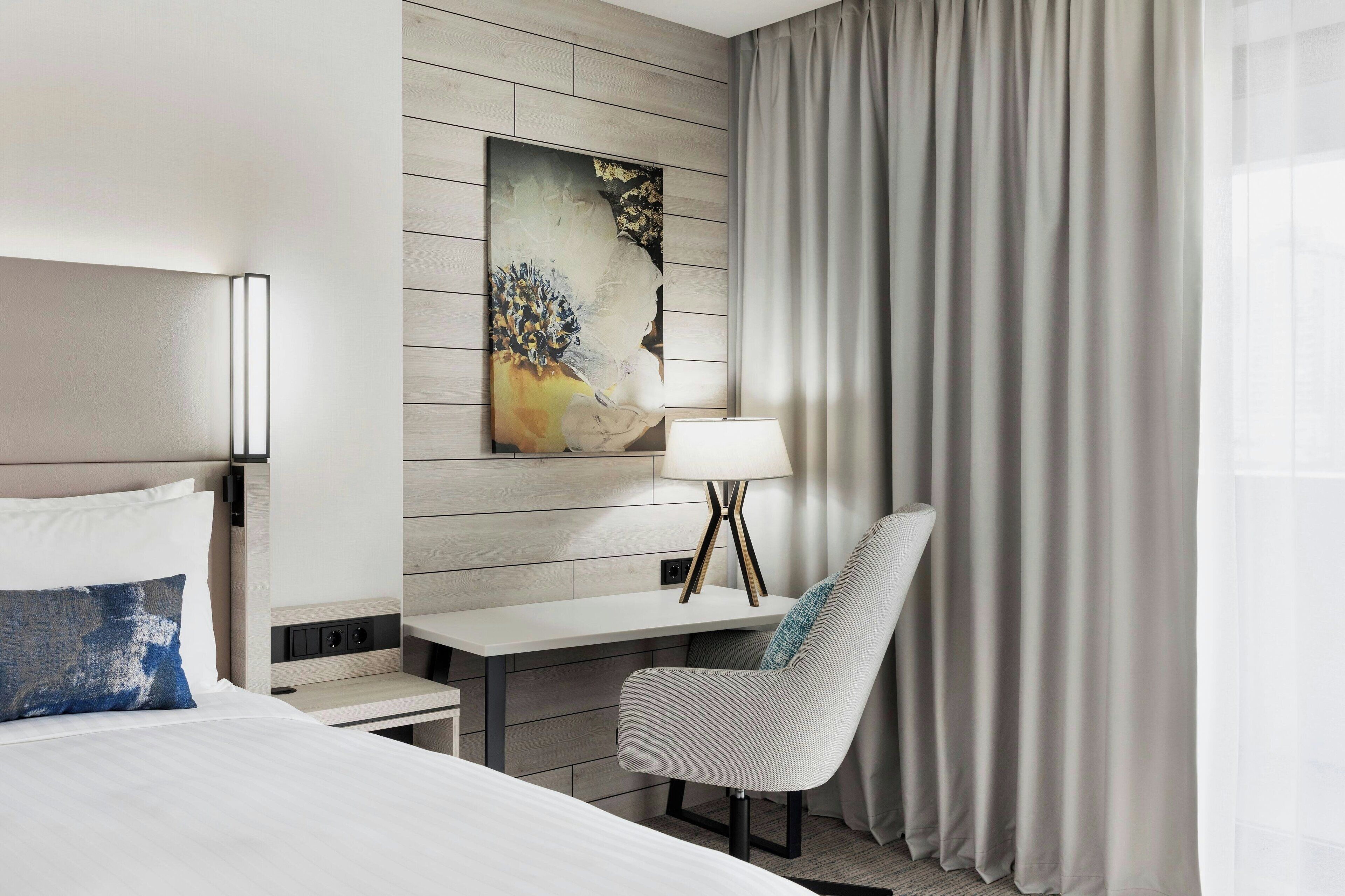 A hotel room with a bed, a desk, a chair, a painting on the wall, and a window with curtains.
