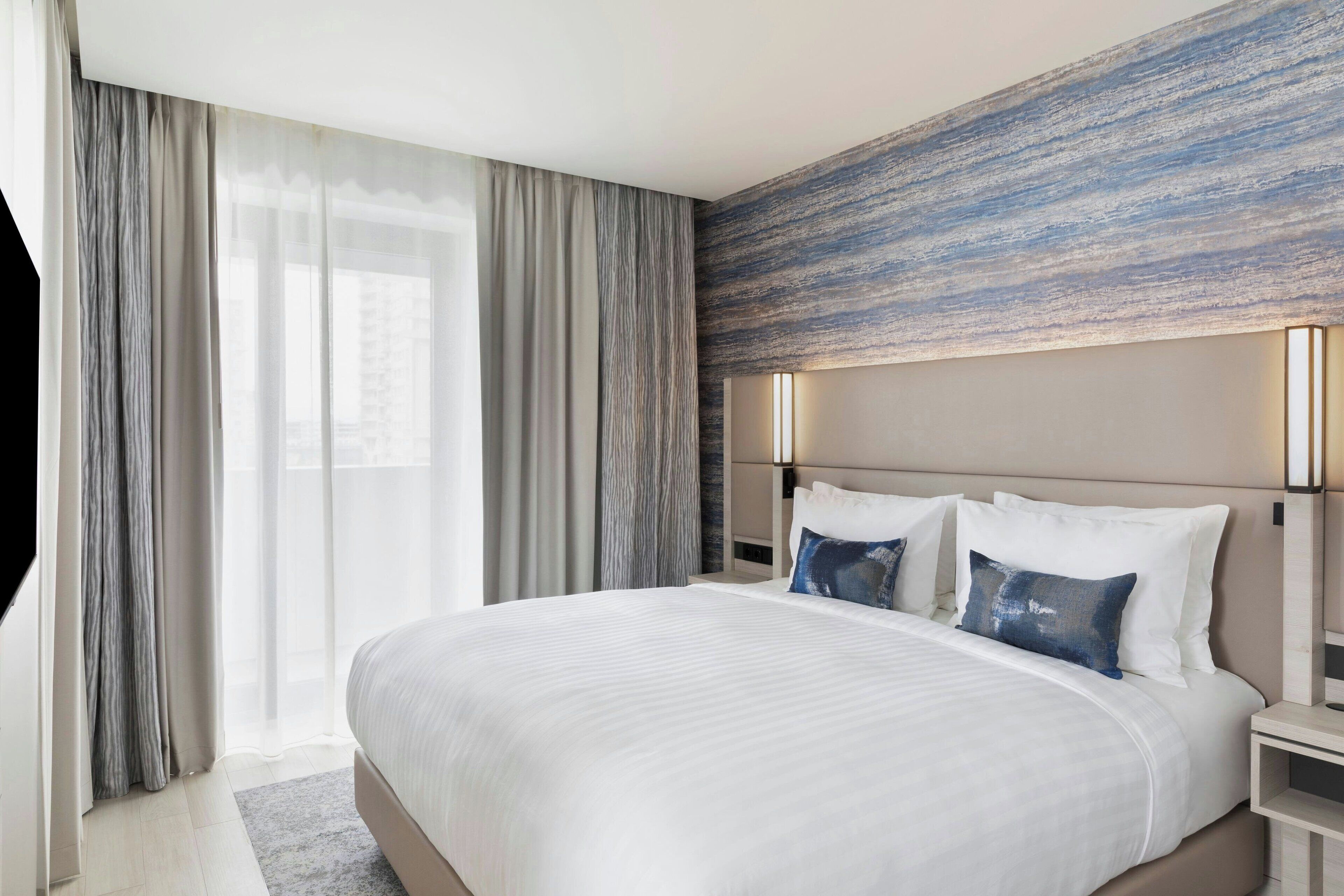 Una habitación de hotel moderna con una cama grande, dos mesitas de noche y vistas a la ciudad a través de cortinas traslúcidas.