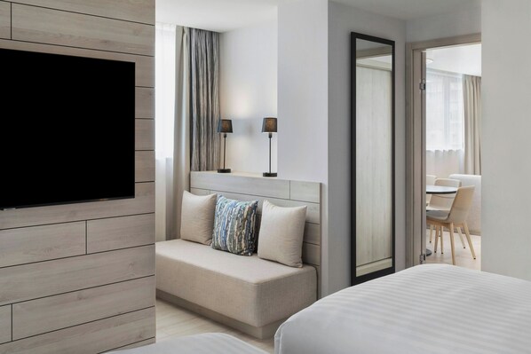 Una habitación de hotel moderna con un televisor de pantalla plana montado en una pared de paneles de madera, un sofá con un cojín estampado, una mesita de noche con lámparas y un espejo grande.