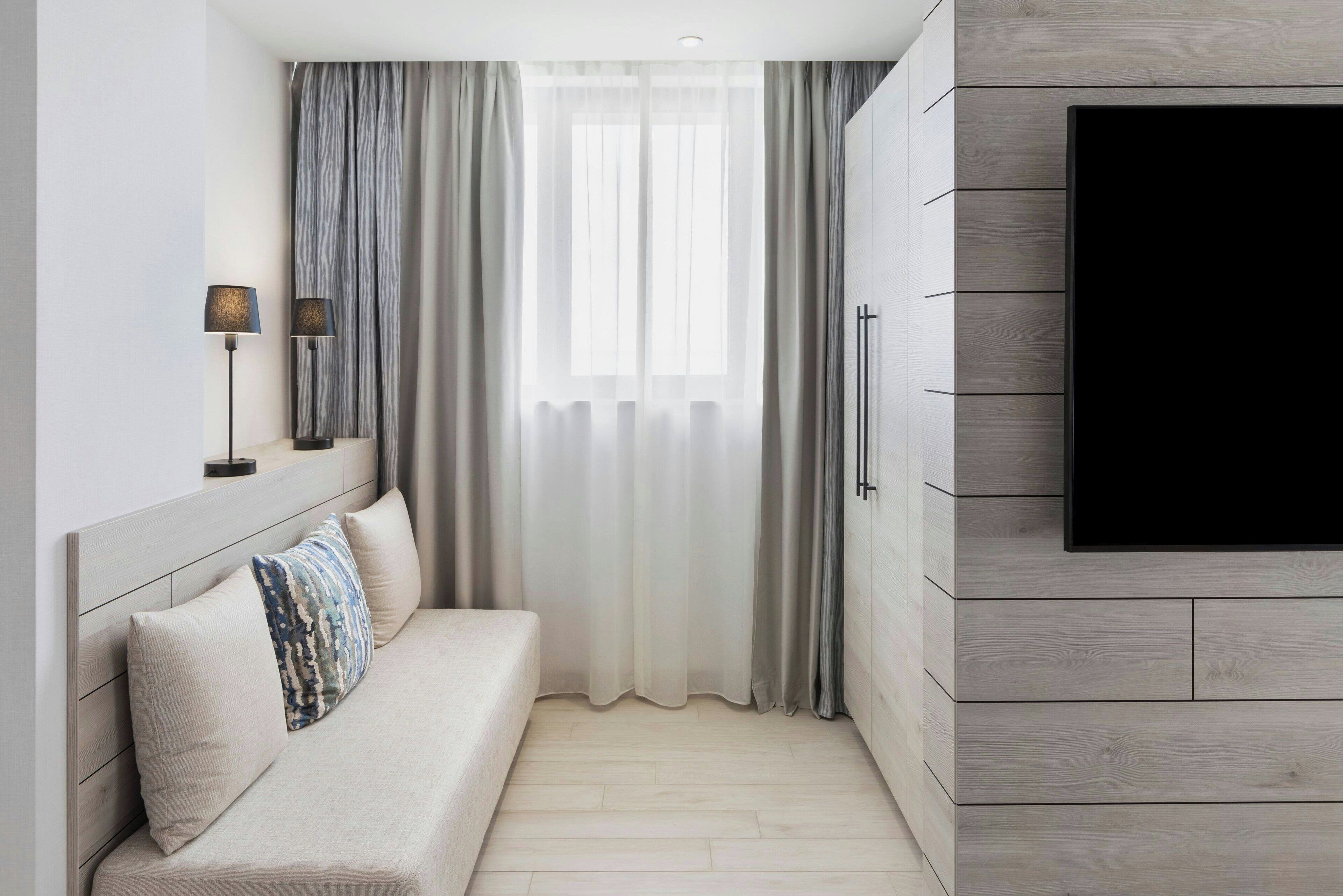 A modern hotel room with a sofa, a TV mounted on the wall, and a large window with sheer curtains.