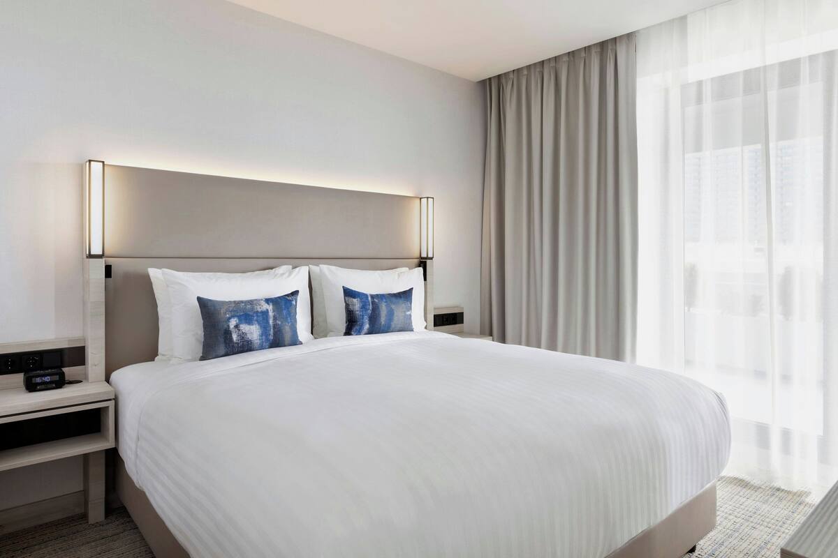 A modern hotel room with a large bed, a bedside table with a clock, and a window with sheer curtains.