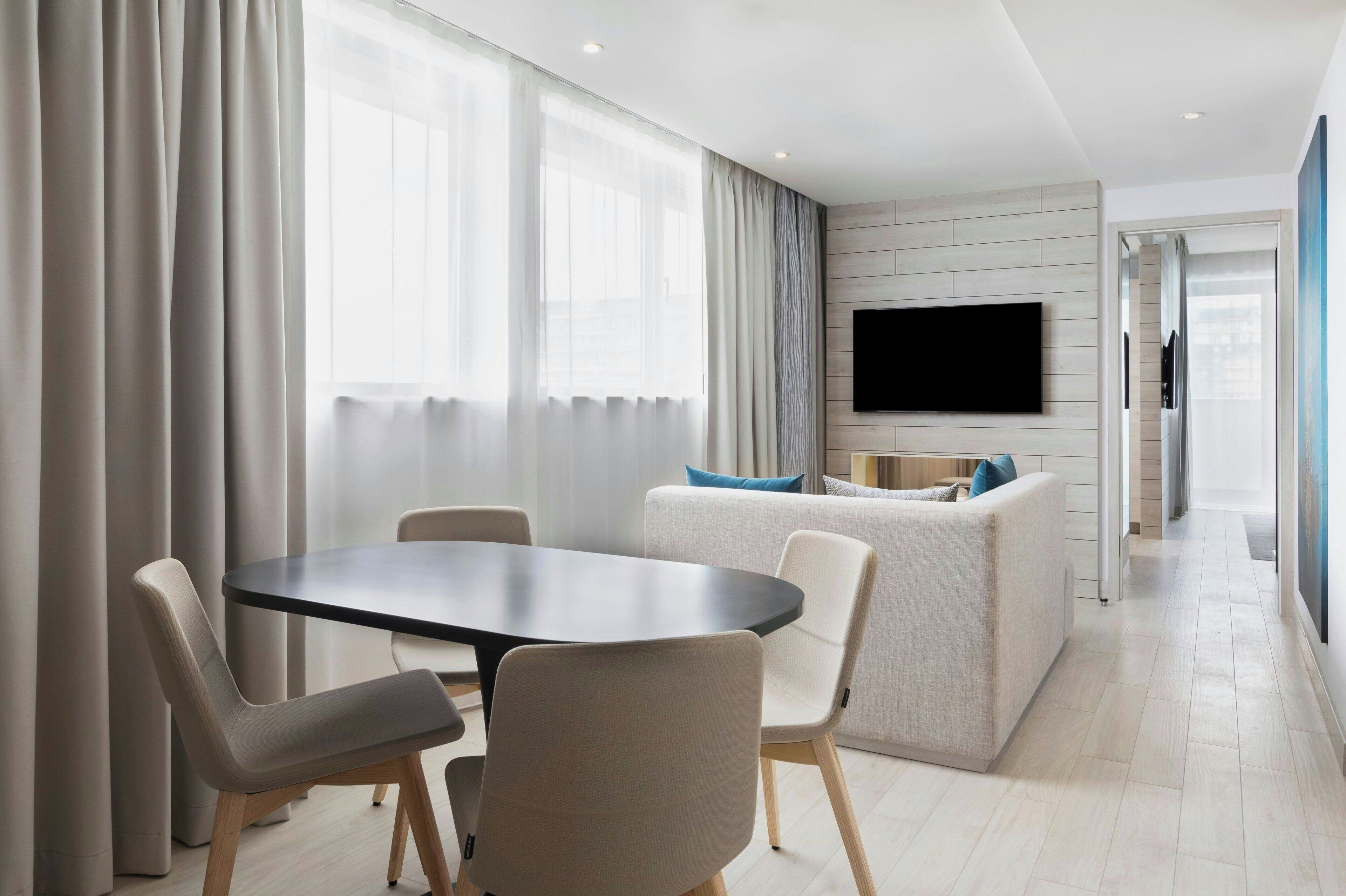 A modern living room with a round table and chairs, a sofa with cushions, a flat-screen TV mounted on the wall, and a large window with curtains.