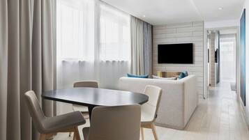 A modern living room with a round table and chairs, a sofa with cushions, a flat-screen TV mounted on the wall, and a large window with curtains.