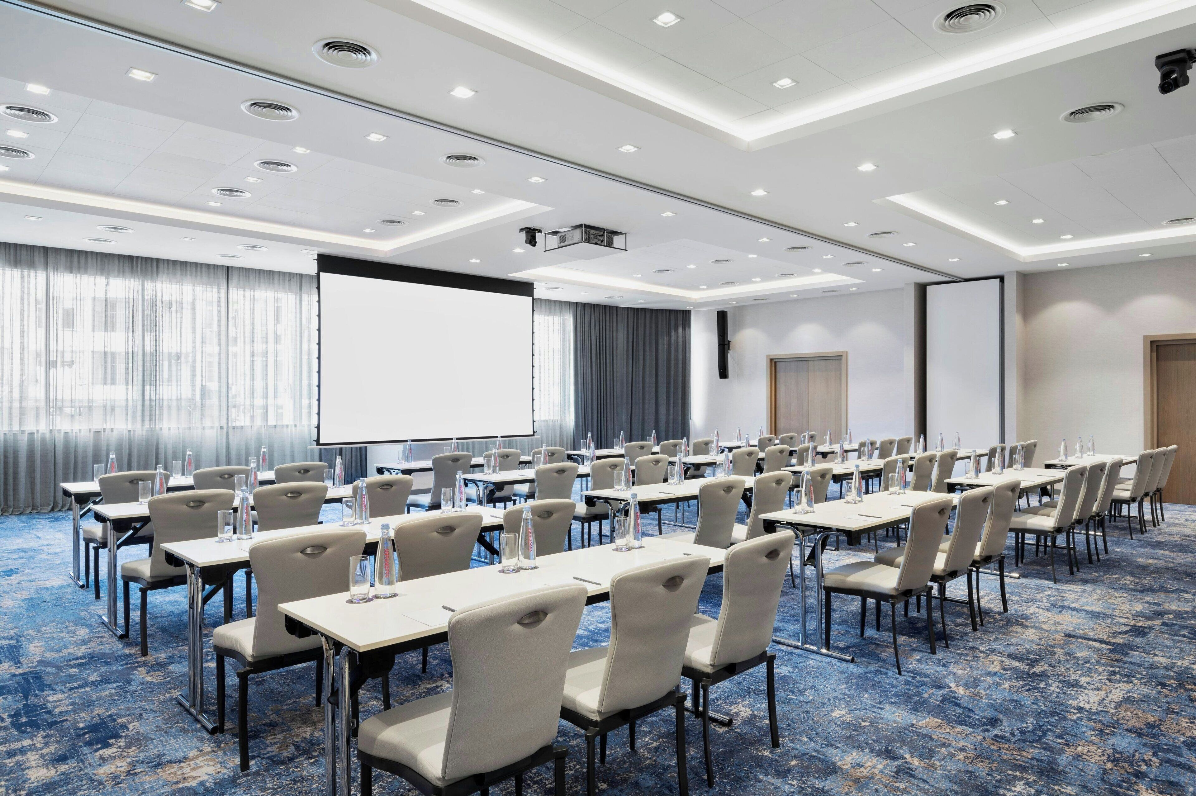 A conference room with rows of tables and chairs, a projector screen, and a large screen on the wall.