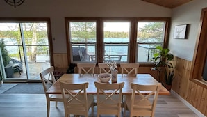 Uma área de refeições com mesa e cadeiras de madeira, grandes janelas com vista para um cais e árvores, e um vaso de planta à direita.