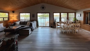 Uma sala de estar ampla com pé direito alto, vigas de madeira, uma grande janela com vista para a água e uma mesa de jantar com cadeiras, além de um canto aconchegante com sofá e mesa de centro.