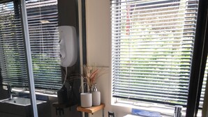 Un baño pequeño con ducha, inodoro y lavamanos. Cuenta con una ventana con persianas, un estante con artículos de aseo y un porta toallas de papel de metal.