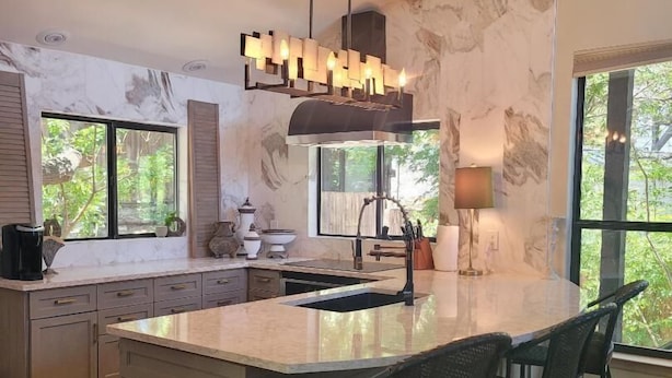 A modern kitchen with a large island, pendant lighting, and a view of greenery outside.