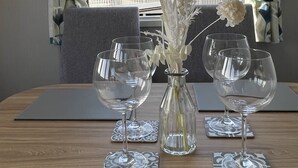 Une table avec deux verres à vin et un vase contenant des fleurs séchées, le tout placé devant une fenêtre ornée de rideaux à motifs floraux.