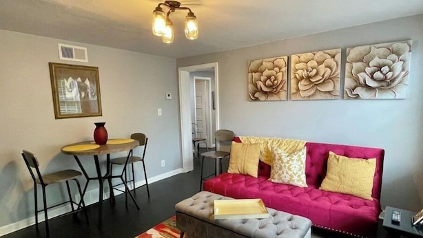 Een woonkamer met een rode bank, een grijze poef, een eettafel met stoelen en een kleurrijk vloerkleed.