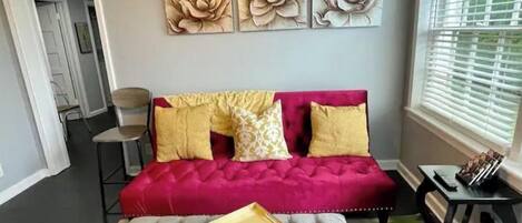 Una habitación con un sofá rojo, un otoman gris y tres cuadros florales colgados en la pared.