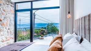Un dormitorio con un ventanal grande, una cama con ropa de cama blanca y marrón, y vistas a un balcón con barandilla y vegetación.