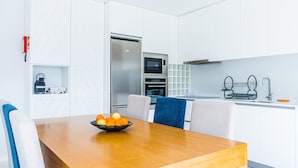 Una cocina moderna con armarios blancos, un refrigerador de acero inoxidable, un horno y un microondas. Hay un comedor de madera con un bol de fruta.