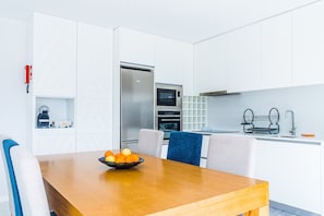 A modern kitchen with white cabinets, a stainless steel refrigerator, an oven, and a microwave. There is a wooden dining table with a bowl of fruit.