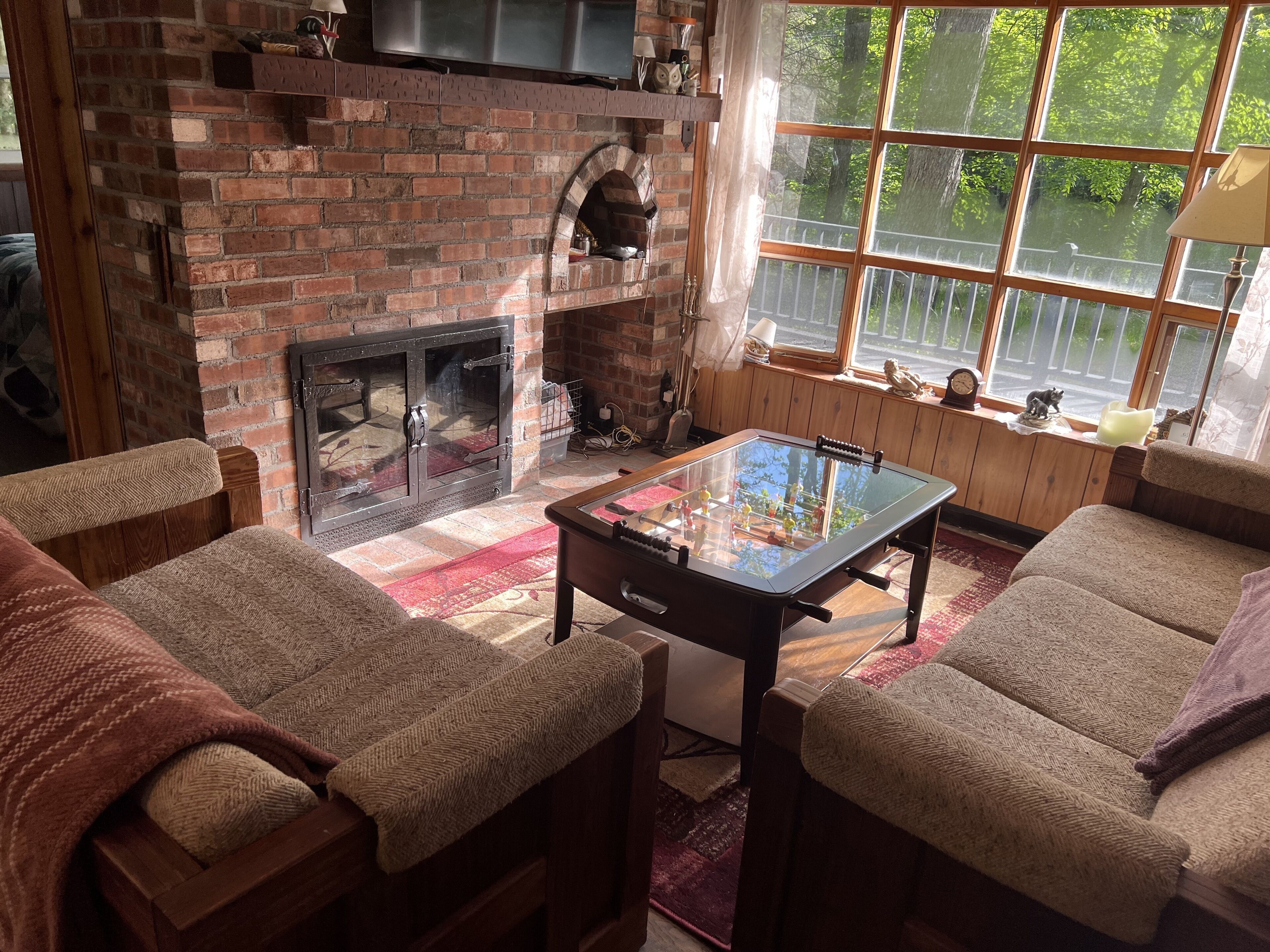 A cozy living room with a brick fireplace, a foosball table, and large windows with a view of trees.