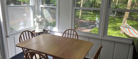 A dining area with a wooden table and chairs, large windows with a view of a garden and hammock.