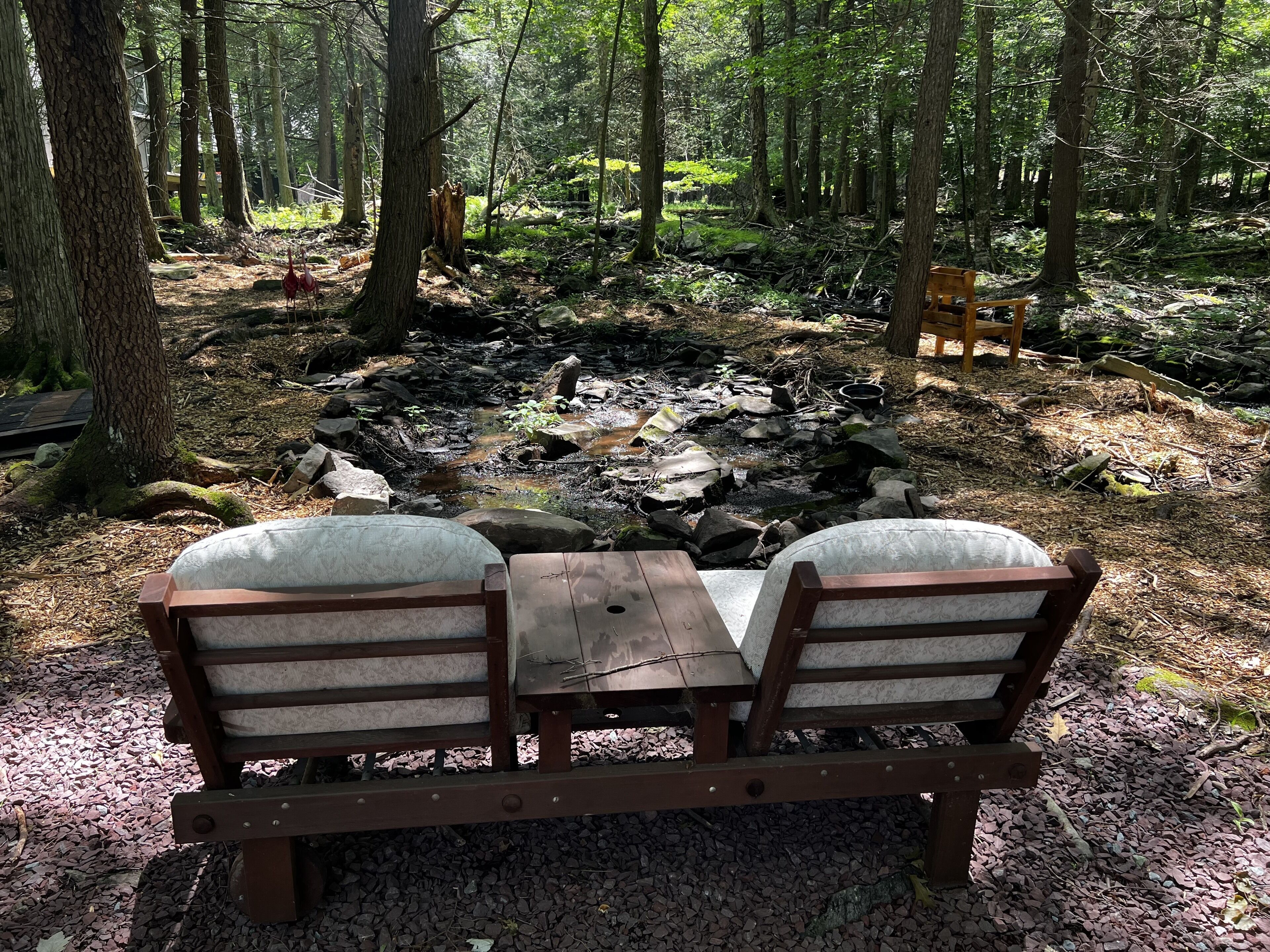 A wooden bench with cushions in a forested area with a small fire pit.