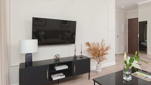 A modern living room with a flat-screen TV mounted on the wall, a black entertainment unit with a lamp, candles, and a vase with pampas grass.
