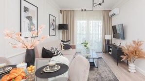 A modern living room with a sofa, a glass coffee table, a TV, and a dining area with pastries and flowers.