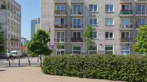 Un edificio residencial de varios pisos con balcones, un sendero pavimentado y un seto en primer plano.
