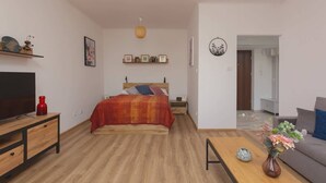 Un dormitorio moderno con una cama de madera, un televisor sobre un mueble de madera, una mesita de noche pequeña y una estantería de pared con objetos decorativos.
