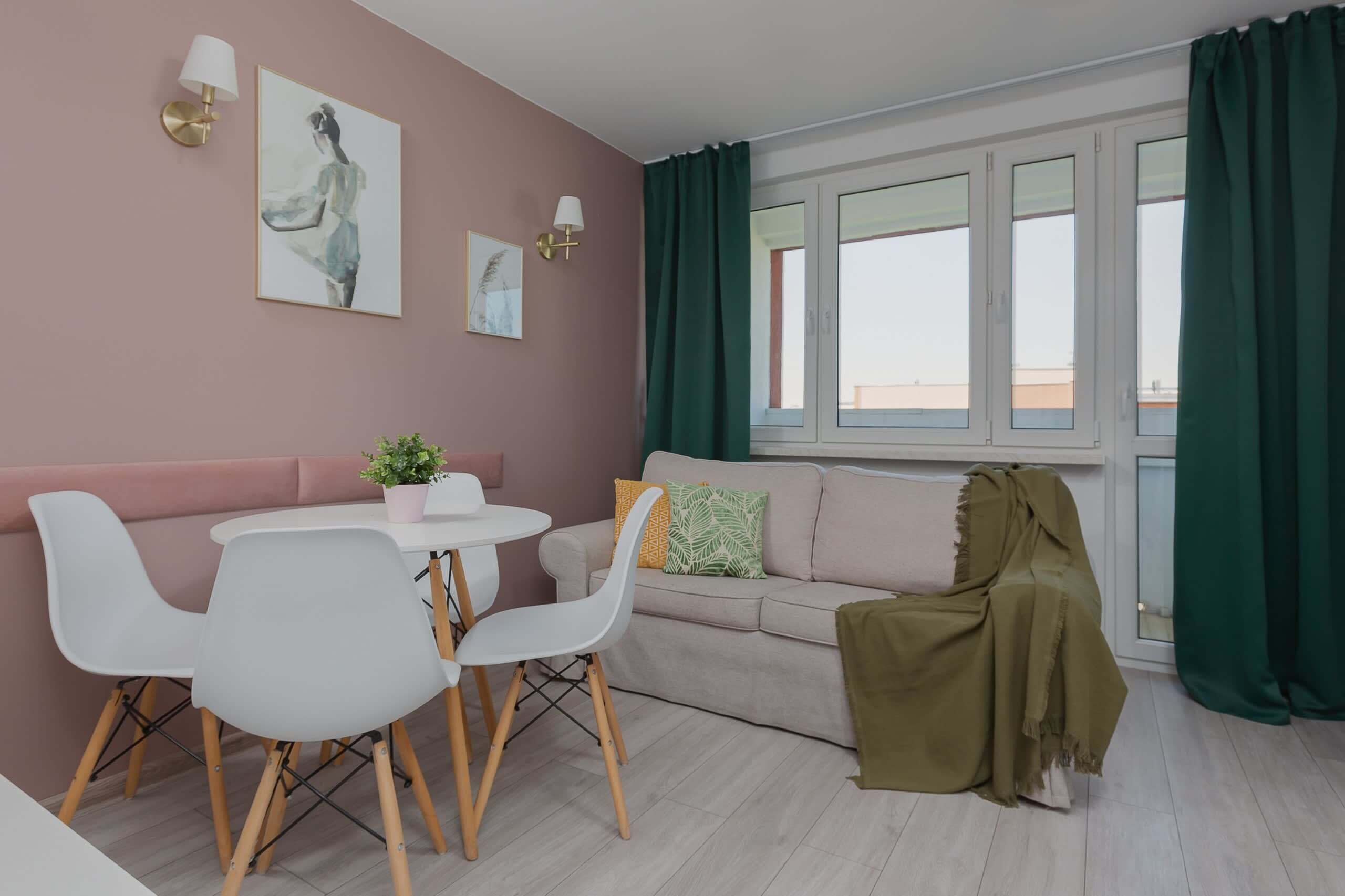 Ein modernes Wohnzimmer mit einem beigen Sofa, einem runden Esstisch mit weißen Stühlen und einem grünen Vorhang, der das Fenster verdeckt.