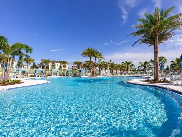 Une grande piscine aux eaux cristallines, entourée de palmiers et de chaises longues, sous un ciel d’un bleu éclatant.