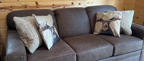 A brown leather sofa with three patterned throw pillows against a wooden wall with a framed deer picture.
