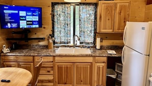 L’intérieur d’une cabane en bois comprend un coin cuisine doté d’un plan de travail en granit, de meubles en bois, d’un évier et d’un réfrigérateur. Un téléviseur est fixé au mur et affiche l’interface d’un service de diffusion en continu.