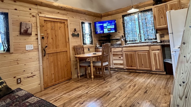 L’intérieur d’une cabane avec des murs en bois, une tête de cerf accrochée au mur, un coin repas avec des chaises, une cuisine avec des meubles en bois et un téléviseur diffusant un service de streaming.