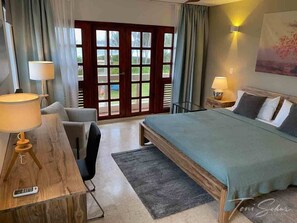 Et hotelværelse med en seng, et skrivebord med lampe, en stol, et lille bord med lampe og et stort vindue med gardiner.