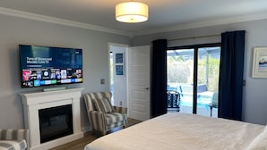 Une chambre d’hôtel avec un grand lit, un téléviseur fixé au-dessus d’une cheminée, un fauteuil à rayures et une vue sur une piscine extérieure, accessible par une porte-fenêtre coulissante.