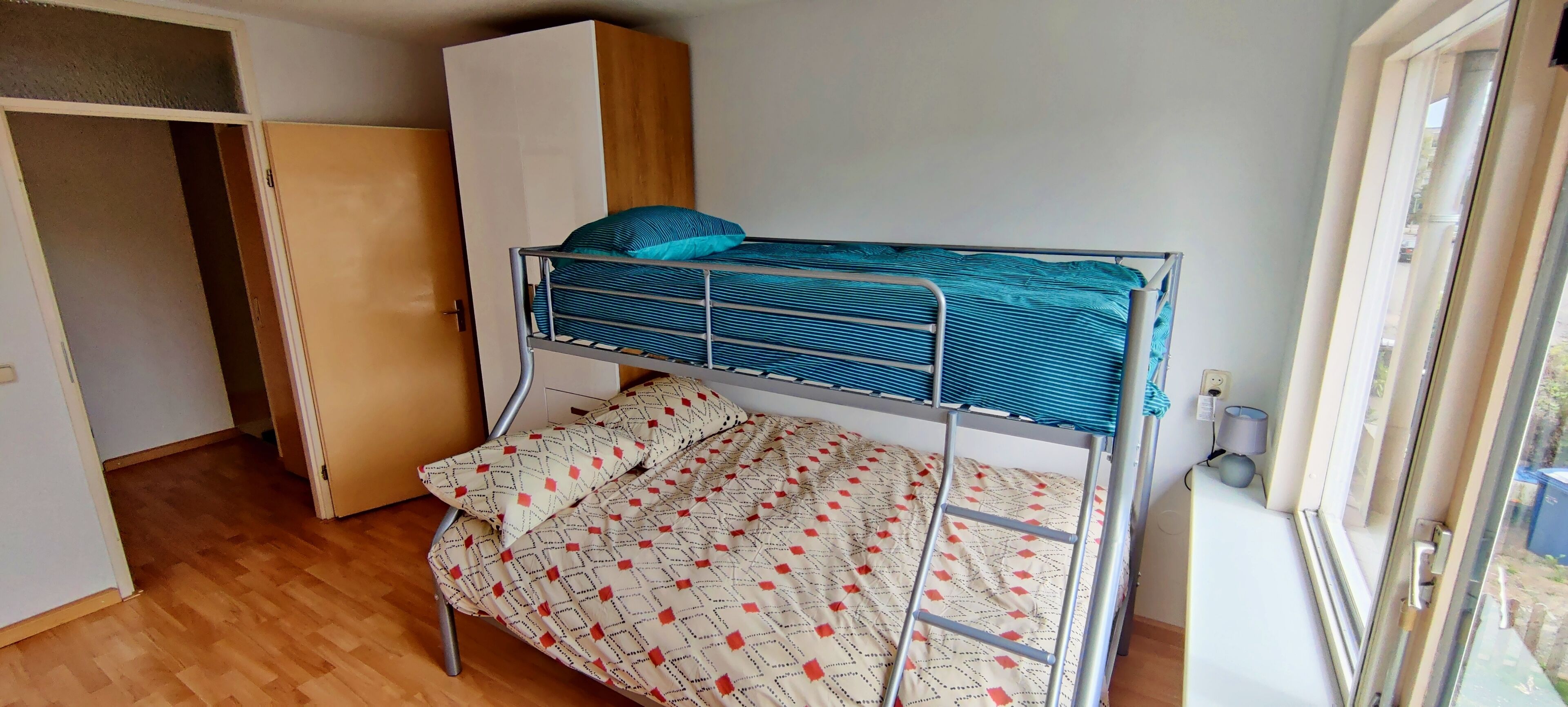 A bunk bed with a patterned blanket, a wardrobe, and a window.