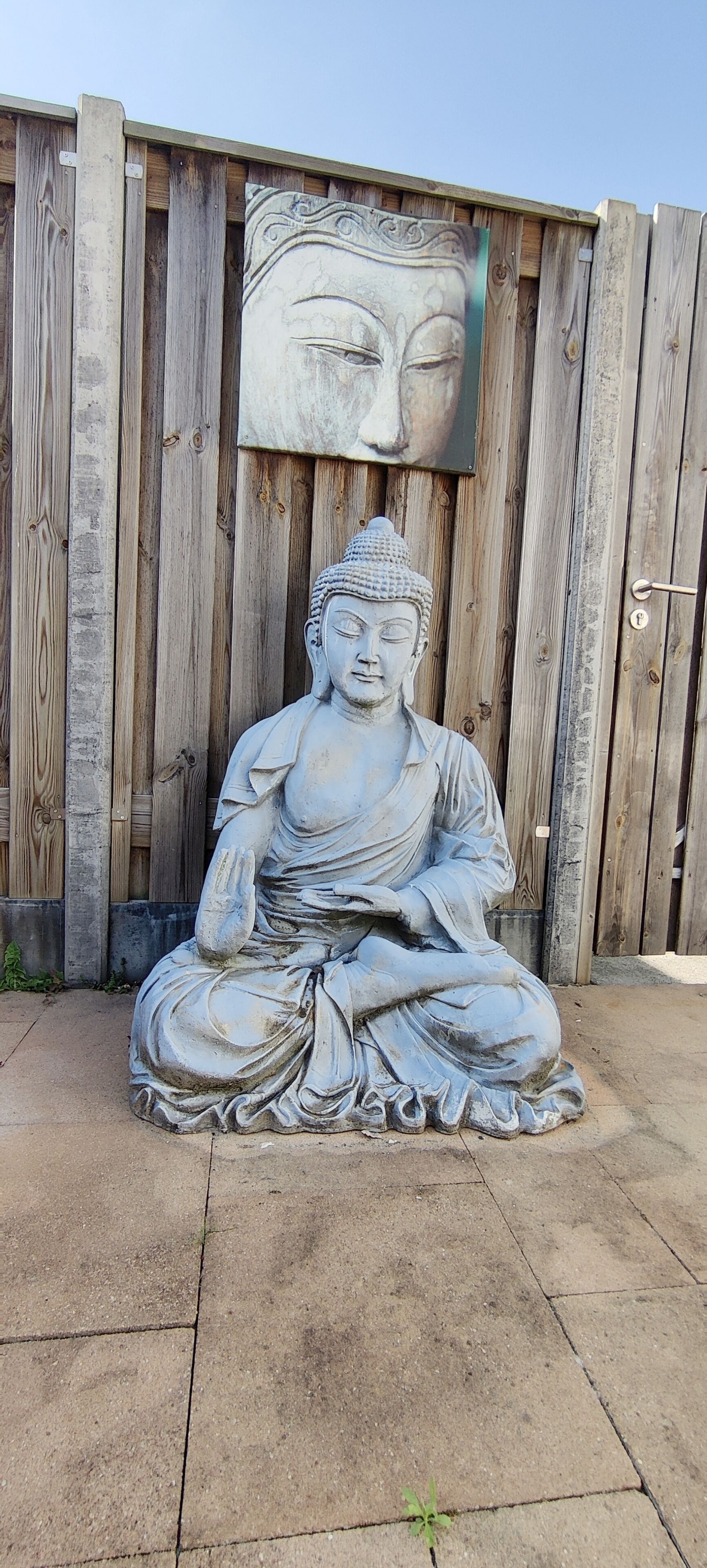A large outdoor statue of a seated figure in a meditative pose, positioned in front of a wooden fence with a large face mural above it.