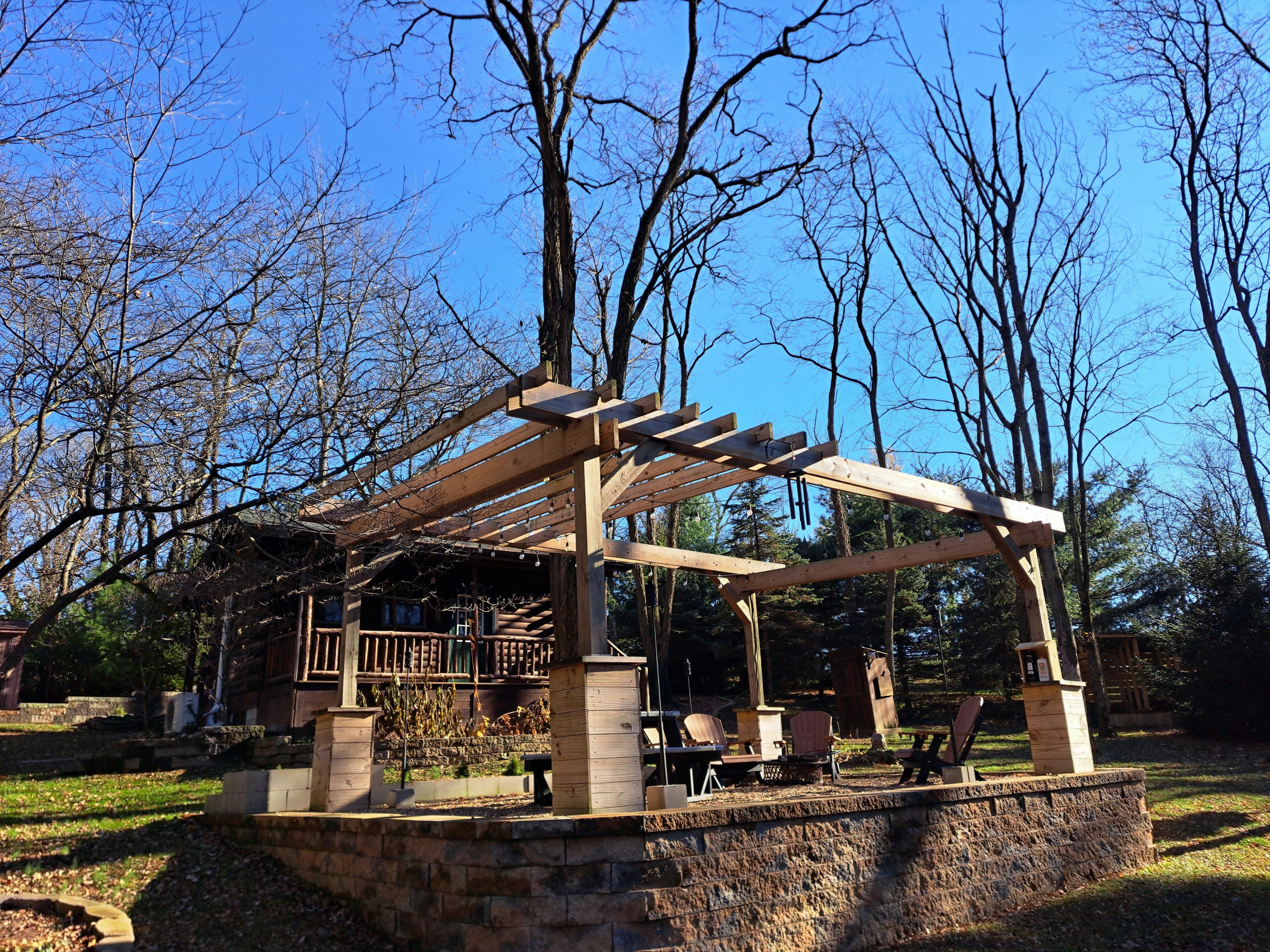 Een houten pergola met zitplaatsen en een stenen basis, omgeven door bomen en een heldere lucht.