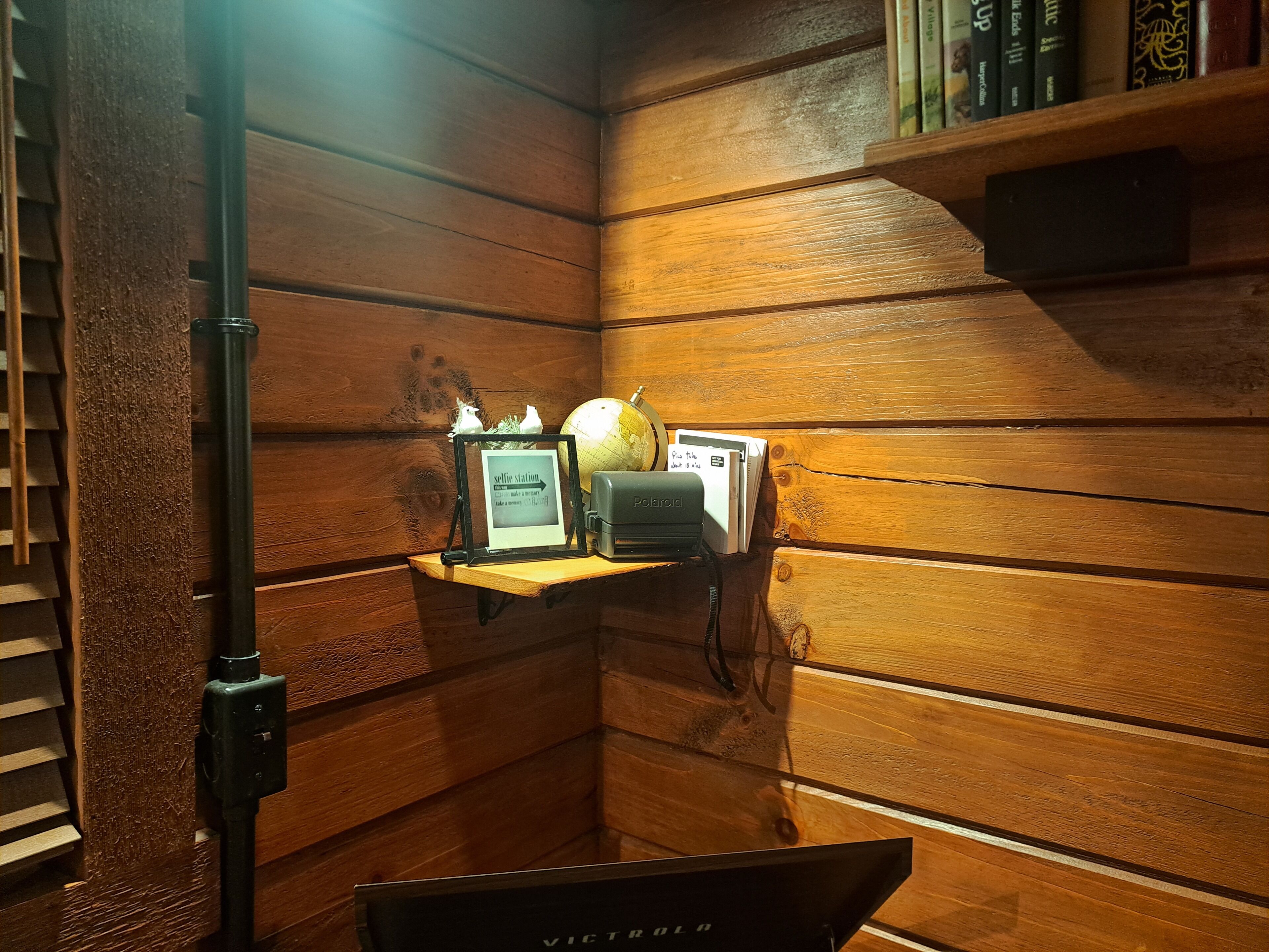 A corner of a room with wooden paneling, a shelf with books, a globe, a framed sign, and a tripod.