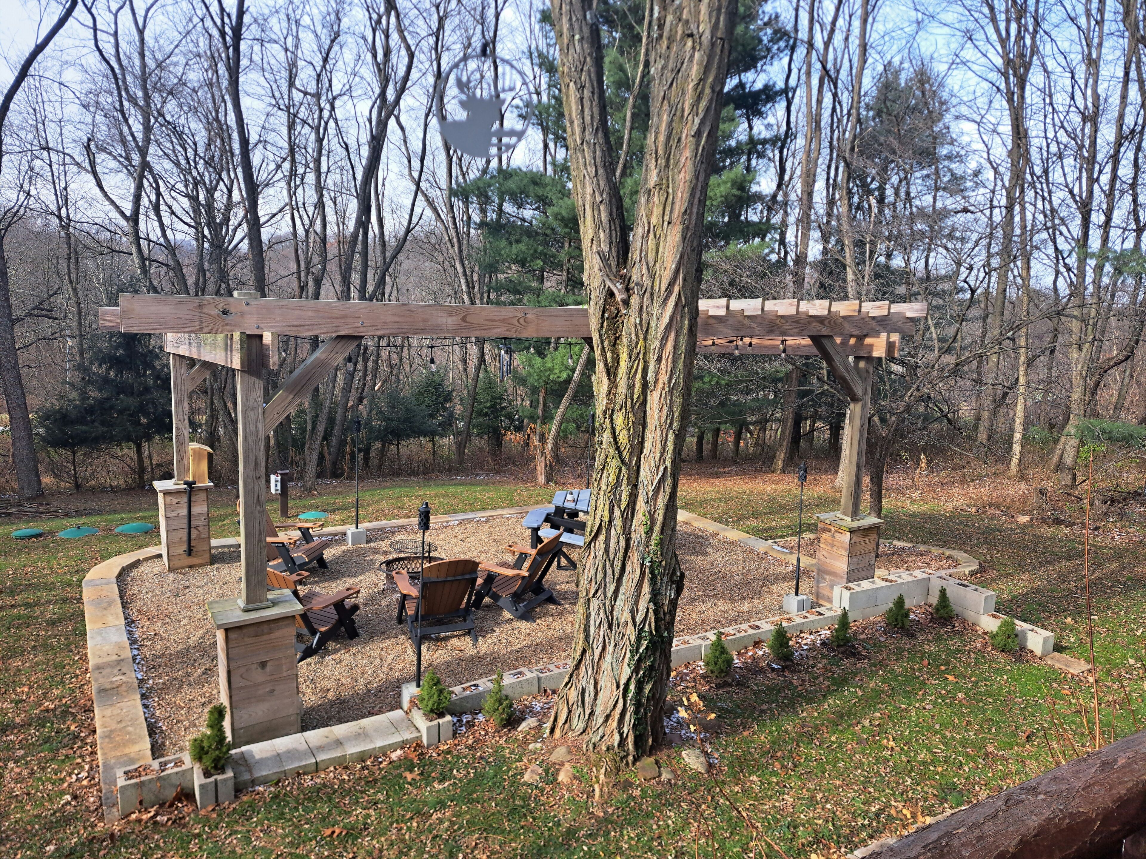 A wooden pergola with seating and a fire pit in a wooded area.