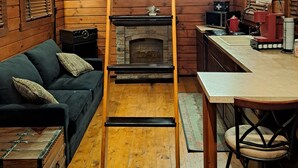 A cozy cabin interior with a wooden ladder leading to a loft, a fireplace, a kitchen area, and a dark-colored sofa.