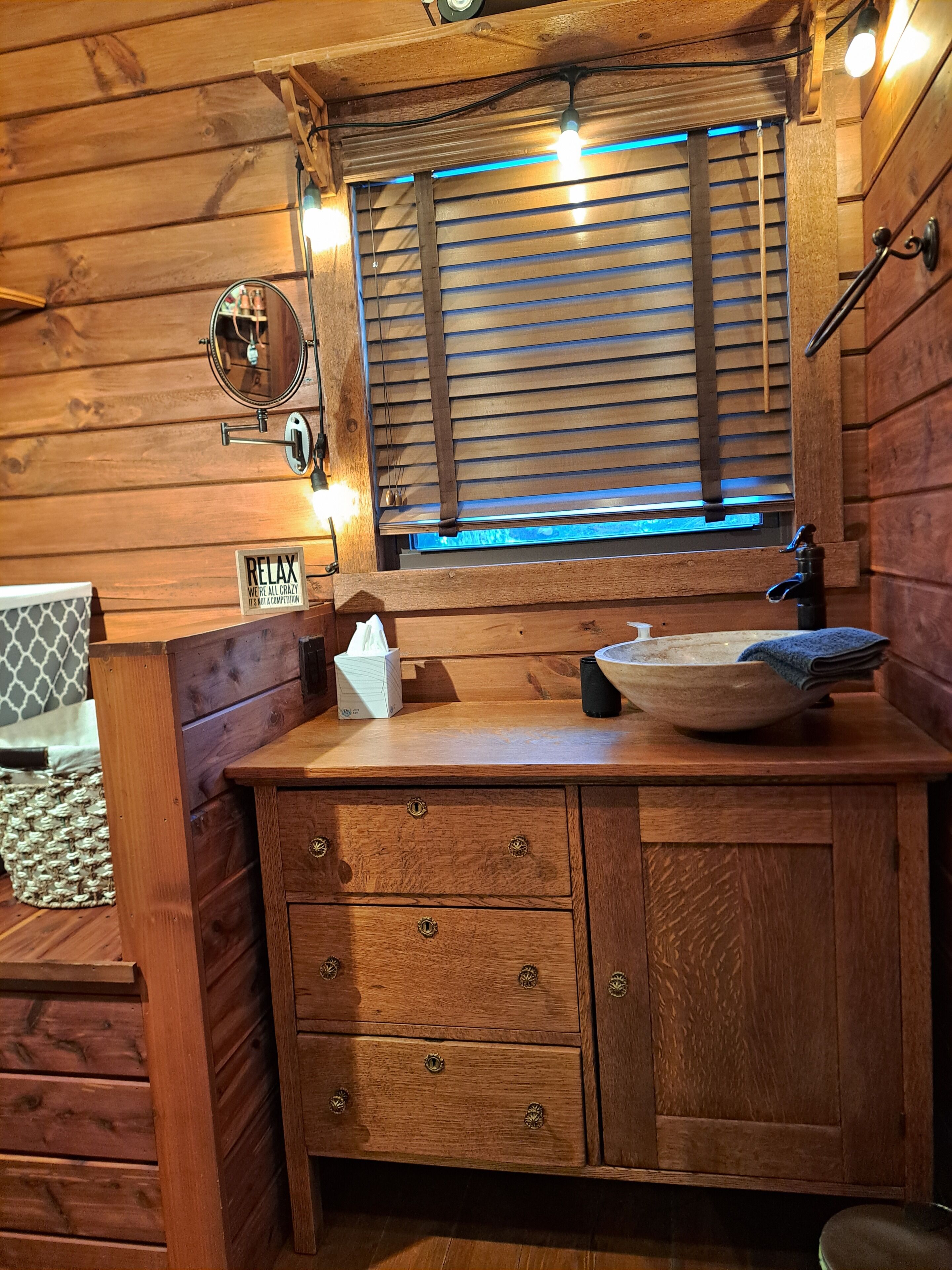 L’intérieur d’un chalet en bois, avec une coiffeuse en bois, un miroir, une fenêtre avec des stores et une pancarte sur laquelle on peut lire « RELAX ».