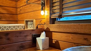 A wooden cabin-style bathroom with a wooden vanity, a mirror, a towel dispenser, and a window with blinds.