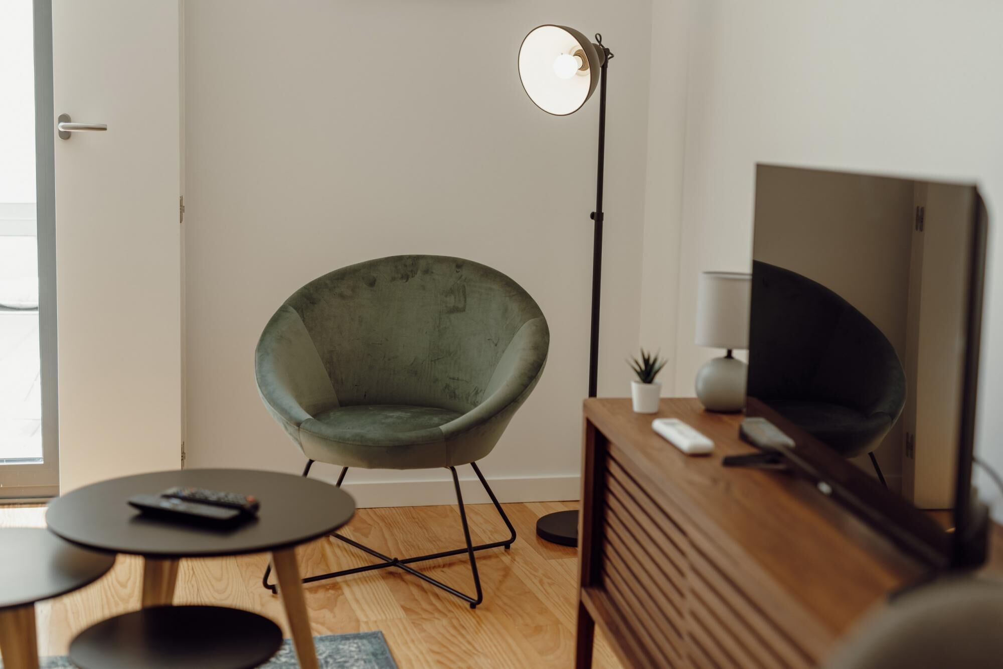 Un salon moderne avec un fauteuil vert, une lampe sur pied, un téléviseur posé sur un meuble en bois et une petite table avec une télécommande.