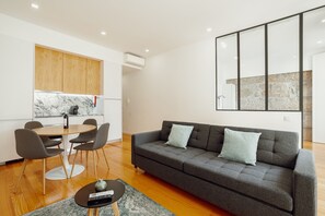 Un salon moderne avec un canapé gris, une table à manger avec des chaises et un coin cuisine doté de meubles en bois et d’un plan de travail en marbre.