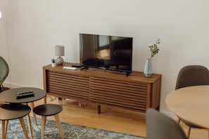 Un salon moderne avec un téléviseur à écran plat posé sur un meuble en bois, une petite table avec une lampe, une plante en pot et deux chaises.