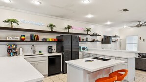 A modern kitchen with white countertops, a central island, and a neon sign above. - Padre Parade • Private Heated Pool • 90-Second Walk to Beach (South Padre Island)