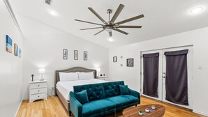 A bedroom with a bed, a teal sofa, a bedside table with a lamp, a ceiling fan, and a sliding door with curtains. - Padre Parade • Private Heated Pool • 90-Second Walk to Beach (South Padre Island)