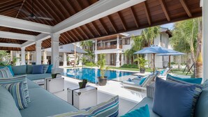 A covered outdoor seating area with a pool, lounge chairs, and a view of a two-story building with balconies. - Villa Windu Asri (Seminyak)