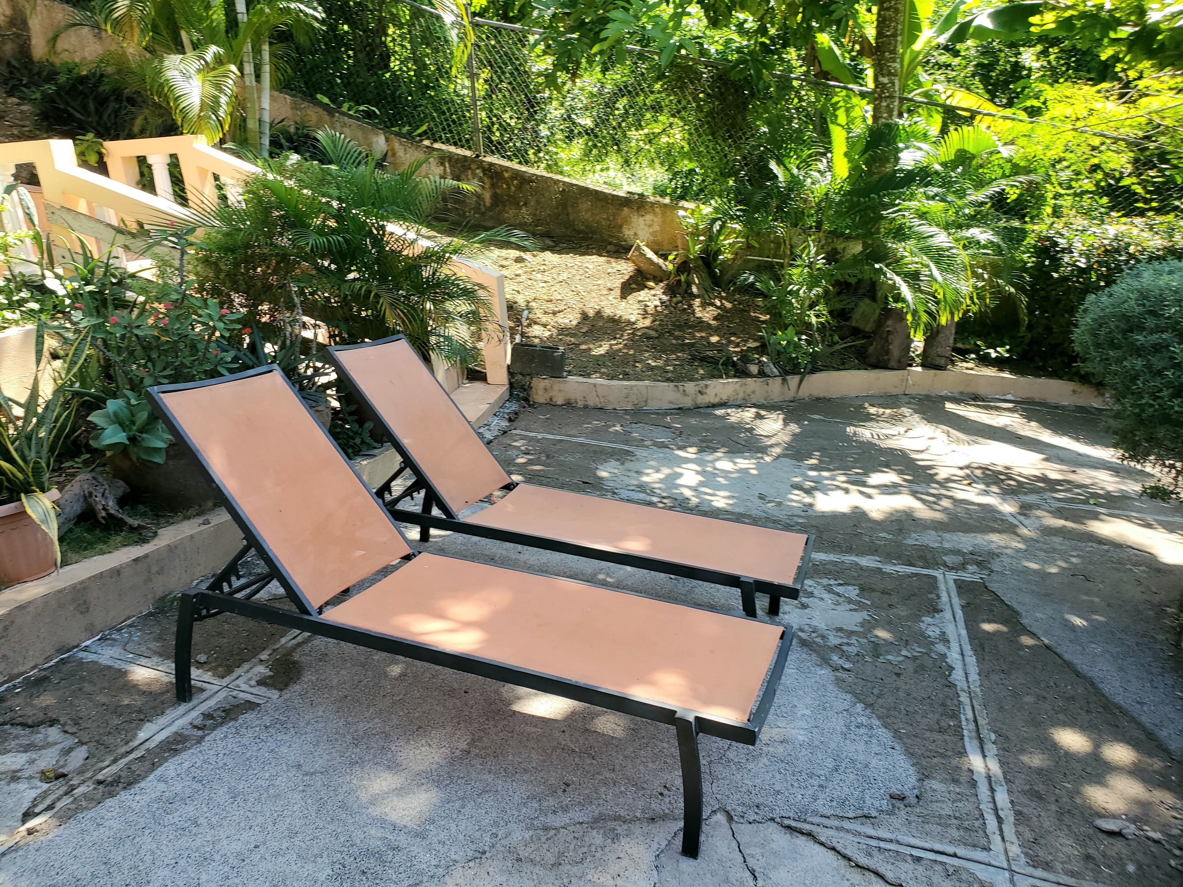 Two sun loungers with metal frames and cushioned seats, positioned on a concrete patio surrounded by lush greenery and a stone wall.