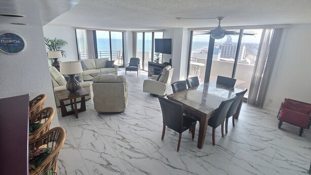 A spacious living area with a marble floor, a dining table, and comfortable seating. Large windows offer a view of the city. - 12th Floor Oceanfront South End Unit overlooking Daytona (Daytona Beach)