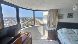 A hotel room with a curved balcony, a bed with floral bedding, a wooden dresser, a nightstand with a lamp, and a view of a coastal cityscape. - 12th Floor Oceanfront South End Unit overlooking Daytona (Daytona Beach)